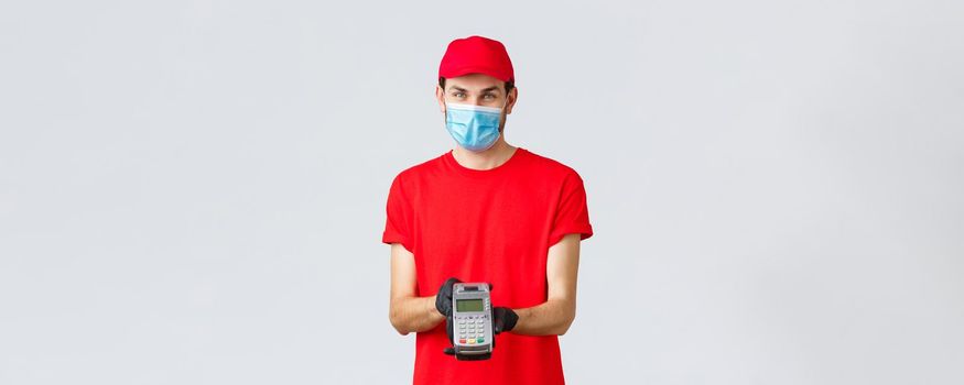 Contactless delivery, payment and online shopping during covid-19, self-quarantine. Pleasant delivery man in face mask and gloves, wear red uniform, giving paying terminal POS to pay for order.