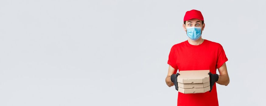 Food delivery, application, online grocery, contactless shopping and covid-19 concept. Surprised courier in red uniform, face mask and gloves, look imressed, bring clients pizza, hold boxes.