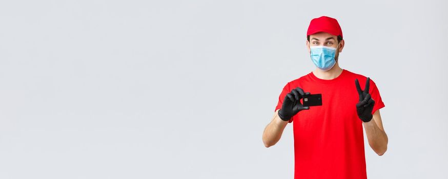 Contactless delivery, payment and online shopping during covid-19, self-quarantine. Friendly courier in red uniform, gloves and face mask provide safe paying option, show credit card and peace sign.