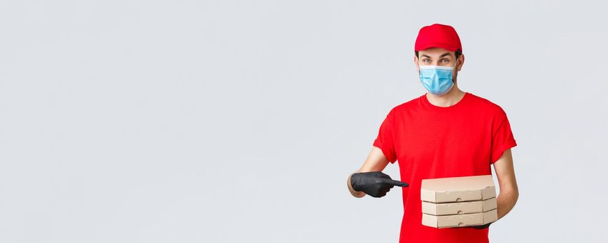 Food delivery, application, online grocery, contactless shopping and covid-19 concept. Cheerful courier in red uniform, face mask and gloves pointing finger at pizza boxes, deliver to client house.
