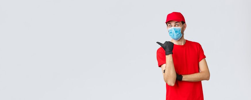 Covid-19, self-quarantine, online shopping and shipping concept. Excited surprised delivery man in red uniform, face mask and gloves, pointing left at promo, showing clients bonuses or application.