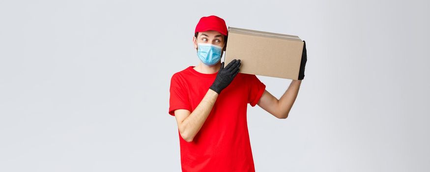 Packages and parcels delivery, covid-19 quarantine delivery, transfer orders. Curious courier in red uniform, gloves and protective face mask, deliver box to client, bring order contactless.