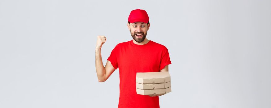 Food delivery, quarantine, stay home and order online concept. Excited and happy courier in red t-shirt and cap, rejoicing holding tasty pizzas, look at boxes and fist pump, grey background.