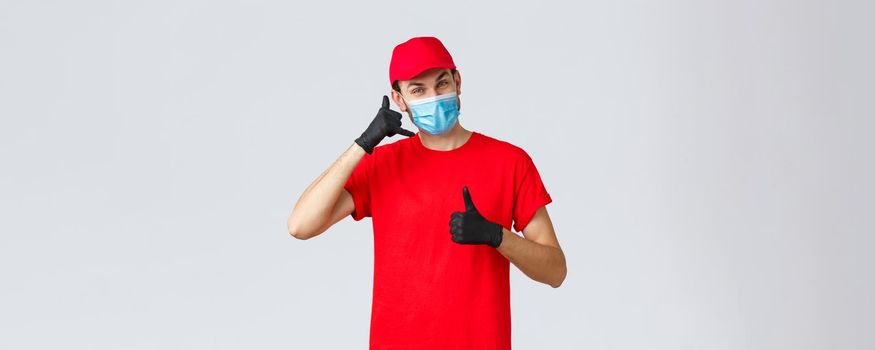Covid-19, self-quarantine, online shopping and shipping concept. Friendly delivery guy in gloves and face mask, carrier uniform, show phone, asking call company order delivery, thumb-up.