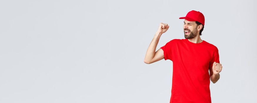 Cheerful and carefree young bearded delivery guy in red uniform cap and t-shirt, singing, yelling yes and fist pump in celebration, triumphing and rejoicing over great news, grey background.