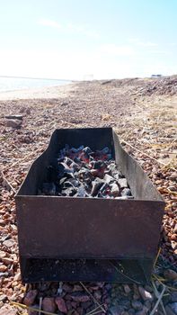 A large barbecue on the shore of the lake, there is smoke from the coals. Bright sunny day, blue lake water, yellow sand with dry branches. Preparation of coals for meat. Food in nature. Lake Balkhash