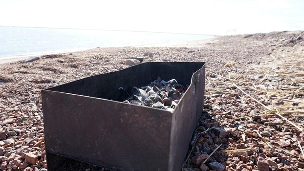 A large barbecue on the shore of the lake, there is smoke from the coals. Bright sunny day, blue lake water, yellow sand with dry branches. Preparation of coals for meat. Food in nature. Lake Balkhash