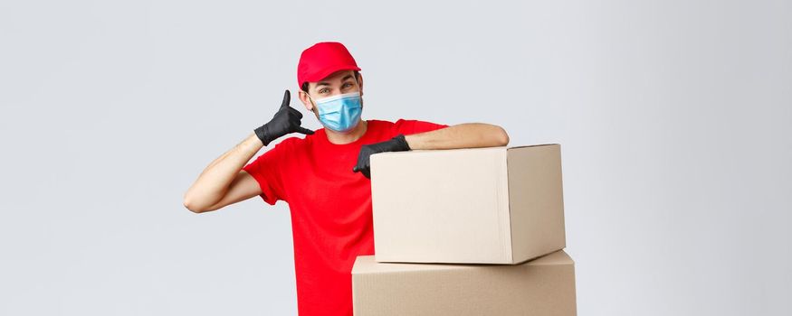 Packages and parcels delivery, covid-19 quarantine and transfer orders. Friendly courier in red uniform, face mask and gloves asking customer give call, show phone sign, stand near boxes.