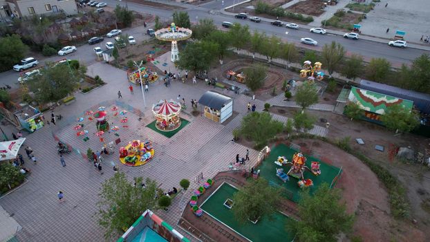 A bird's-eye view of children's attractions from a drone. Bright lights from different lanterns. Carousels are spinning. People are resting. The attractions are located on the shore of Lake Balkhash