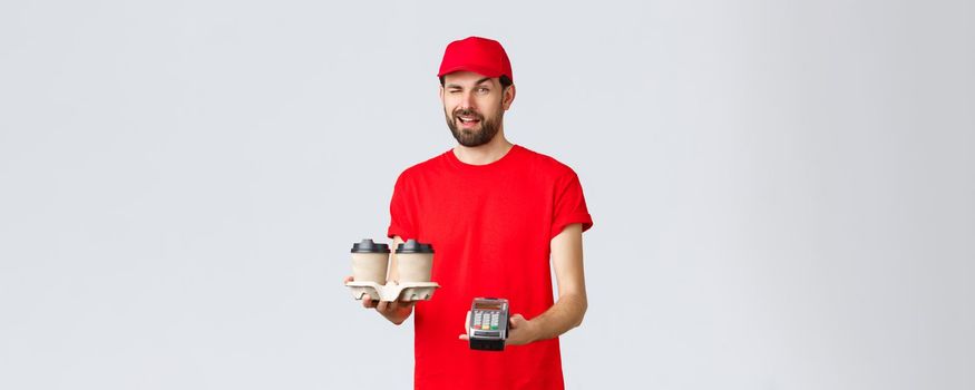 Food delivery, quarantine, stay home and order online concept. Cheeky courier in red uniform cap and t-shirt, wink to client as handing POS terminal and coffee deliver to pay contactless.