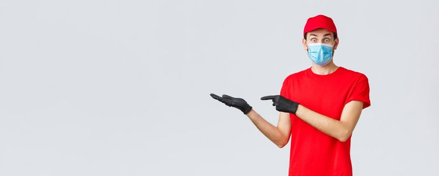 Groceries and packages delivery, covid-19, quarantine and shopping concept. Surprised and excited courier in red uniform, face mask and gloves introduce good new bonus offer, promo, showing banner.