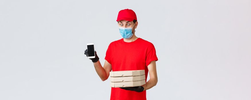 Food delivery, application, online grocery, contactless shopping and covid-19 concept. Amused funny courier in red uniform, face mask and gloves, showing smartphone screen app and hold pizza boxes.