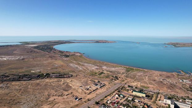 Drone view of Bertys Bay, Lake Balkhash. The blue-green water of the lake reflects the sky. There is a town and a factory on the shore. The coastal line is clearly visible, low houses