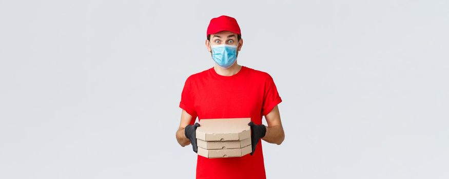 Food delivery, application, online grocery, contactless shopping and covid-19 concept. Surprised courier in red uniform, face mask and gloves, look imressed, bring clients pizza, hold boxes.