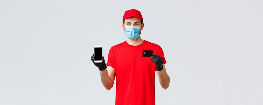 Contactless delivery, payment and online shopping during covid-19, self-quarantine. Smiling courier in red uniform, gloves and face mask showing smartphone screen and credit card, app for orders.