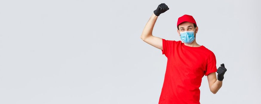 Groceries and packages delivery, covid-19, quarantine and shopping concept. Cheerful courier red uniform, gloves and face mask, raising hands up, chanting rejoicing over good news, fight coronavirus.