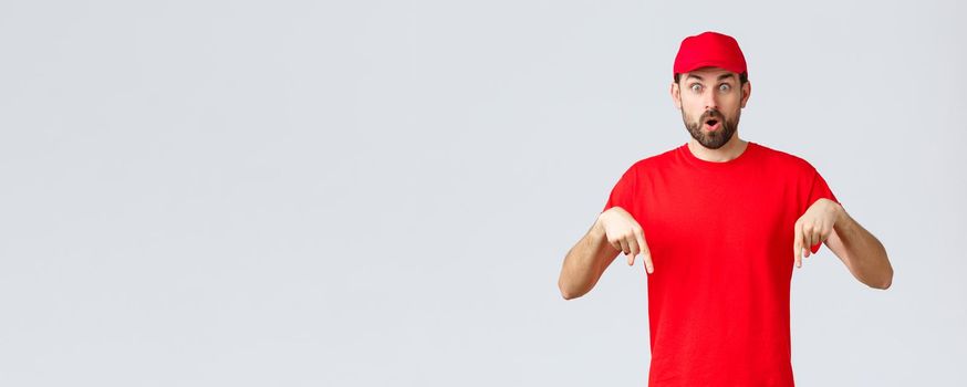 Online shopping, delivery during quarantine and takeaway concept. Surprised and intrigued courier inform clients, employee in red uniform cap and t-shirt pointing fingers down, say wow.