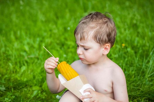 A cute blond boy appetizingly eats corn in the summer, sitting on the bank of the river on the juicy grass. Funny facial expression. summer heat, a child without a t-shirt