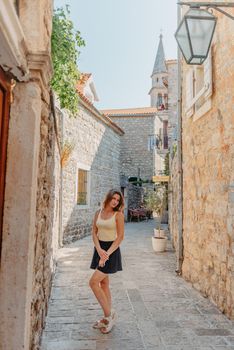 Girl tourist walking through ancient narrow street on a beautiful summer day in MEDITERRANEAN MEDIEVAL CITY , OLD TOWN bUDVA, MONTENEGRO. Young beautiful cheerful woman walking on old street at tropical town. Pretty girl looking at you and smiling