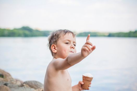 A cute blond boy appetizingly eats ice cream in the summer, sitting on the bank of the river. Cool off by the water. Funny facial expression. summer heat