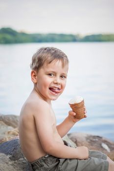 A cute blond boy appetizingly eats ice cream in the summer, sitting on the bank of the river. Cool off by the water. Funny facial expression. summer heat