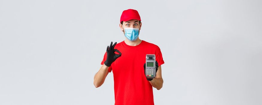 Contactless delivery, payment and online shopping during covid-19, self-quarantine. Excited courier in red uniform, face mask and gloves recommend pay order with POS terminal and credit card.