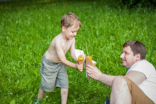 A cute blond boy and his father eat corn in the summer, sitting on the bank of the river on the juicy grass. Funny facial expression. summer heat, a child without a t-shirt