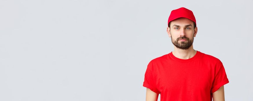 Online shopping, delivery during quarantine and takeaway concept. Young handsome bearded courier in red service uniform with cap and t-shirt, employee working transfering orders.