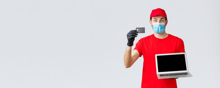 Customer support, covid-19 delivery packages, online orders processing concept. Surprised excited courier in red uniform, face mask and gloves, showing credit card to pay orders and laptop.