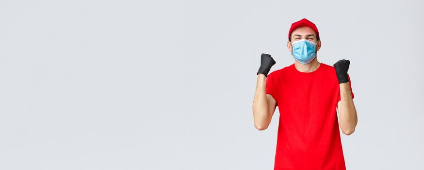 Covid-19, self-quarantine, online shopping and shipping concept. Excited, happy courier in red uniform, medical face mask, fist pump in rejoice. Delivery guy celebrating success, achieve goal.