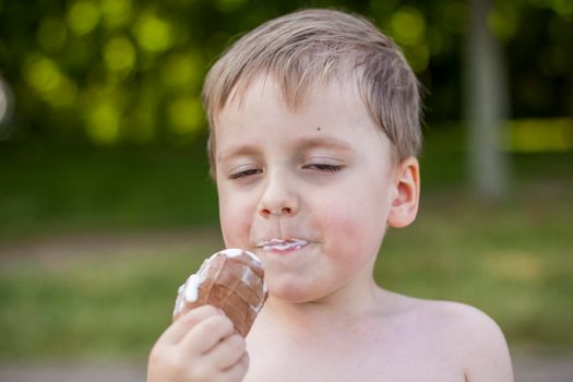 A cute blond boy appetizingly eats ice cream in the summer, sitting on the bank of the river. Cool off by the water. Funny facial expression. summer heat