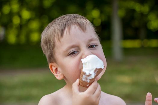 A cute blond boy appetizingly eats ice cream in the summer, sitting on the bank of the river. Cool off by the water. Funny facial expression. summer heat