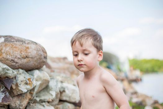 A cute blond boy experiences the joy of walking on a summer day by the river, sitting on the juicy grass. Funny facial expression. summer heat, a child without a T-shirt. Summer