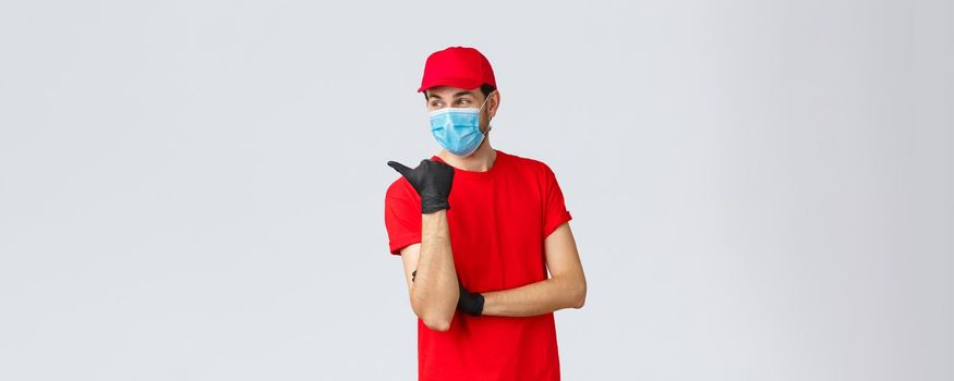 Covid-19, self-quarantine, online shopping and shipping concept. Delivery guy in red uniform, gloves and face mask, pointing left and smiling, recommend client use courier service.