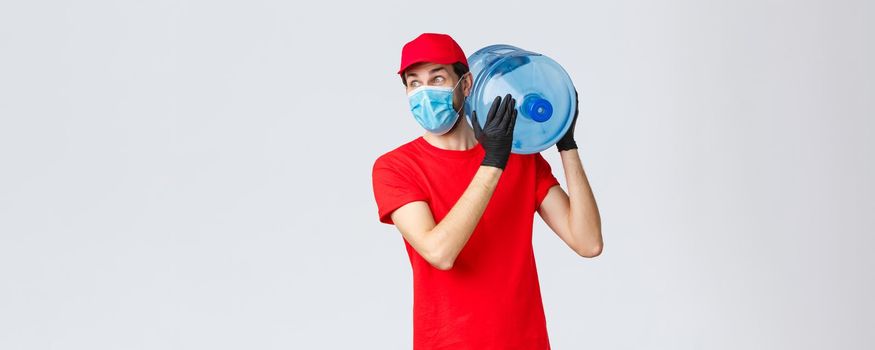 Takeaway, food and groceries delivery, covid-19 contactless orders concept. Excited and impressed courier in red uniform, gloves and face mask, hold bottled water on shoulder, look away astonished.