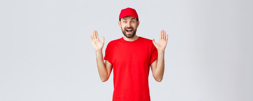 Online shopping, delivery during quarantine and takeaway concept. Happy cheerful courier in red t-shirt and cap, company uniform, hands up surprised and amused, standing grey background.