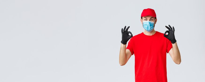 Covid-19, self-quarantine, online shopping and shipping concept. Excited delivery guy in red cap, t-shirt and face mask working coronavirus outbreak, show okay sign, guarantee safe courier service.