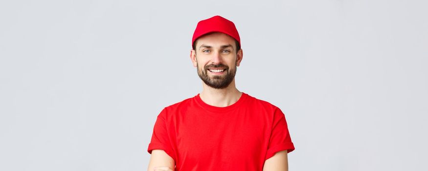 Online shopping, delivery during quarantine and takeaway concept. Handsome bearded courier in red uniform, smiling cheerful, cross arms chest, ready to deliver your order, grey background.