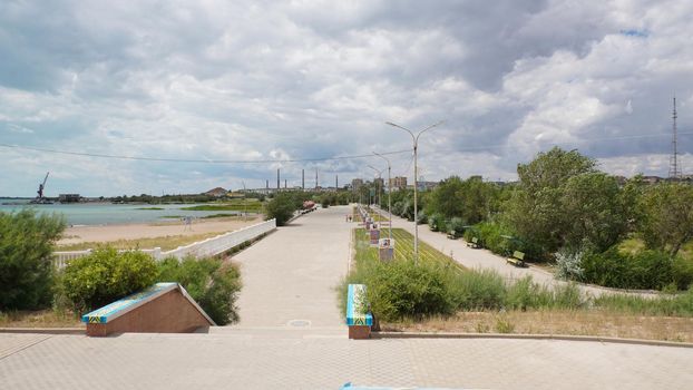 A small town with big clouds and a factory. Alley, green trees swaying in the wind, flowers in flower beds. People are walking. In the distance you can see a lake, reeds grow. Balkhash city