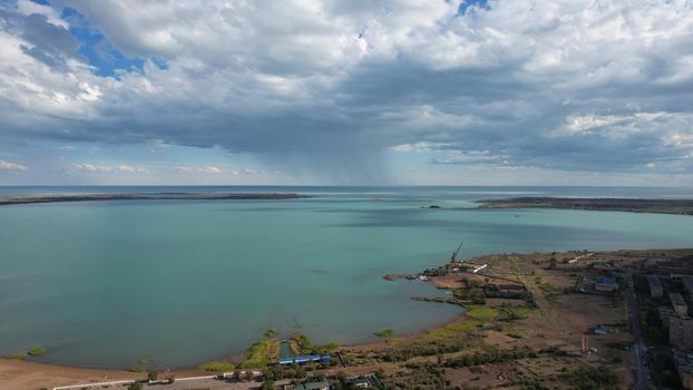 Drone view of Bertys Bay, Lake Balkhash. The blue-green water of the lake reflects the sky and clouds. There is a town and a factory on the shore. The coastal line is clearly visible, low houses