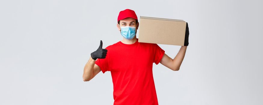 Packages and parcels delivery, covid-19 quarantine delivery, transfer orders. Cheerful courier in red uniform, gloves and face mask, thumb-up, no problem deliver order box to customers.