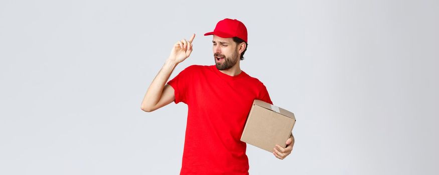 Order delivery, online shopping and package shipping concept. Carefree handsome bearded courier in red uniform, cap and t-shirt, holding box package, singing song, grey background.