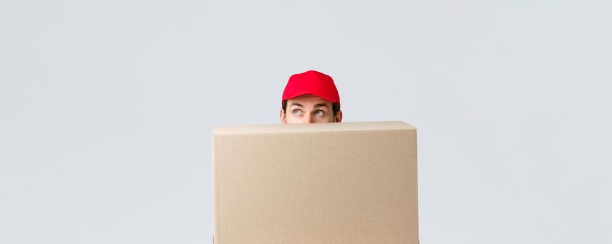 Packages and parcels delivery, covid-19 quarantine and transfer orders. Cute courier in red uniform cap hiding behind large box order, looking up intrigued, peeking over grey background.