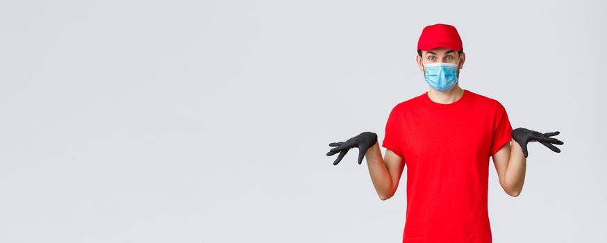 Covid-19, self-quarantine, online shopping and shipping concept. Funny courier in black rubber gloves and face mask, delivery guy in uniform gesturing reluctant, standing grey background.