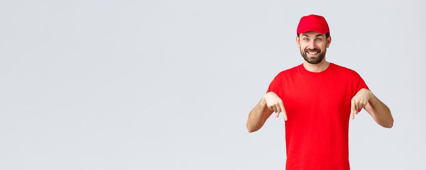 Online shopping, delivery during quarantine and takeaway concept. Cheerful friendly courier in red uniform cap and t-shirt, pointing fingers down, inform clients, recommend advertisement.