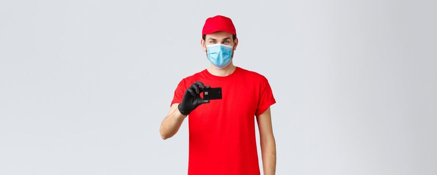 Contactless delivery, payment and online shopping during covid-19, self-quarantine. Handsome courier in red uniform, cap, medical face mask and gloves, show credit card, order internet.
