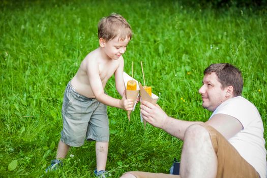 A cute blond boy and his father eat corn in the summer, sitting on the bank of the river on the juicy grass. Funny facial expression. summer heat, a child without a t-shirt