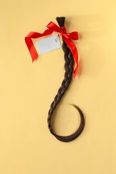 Long brown braid with red ribbon and empty label on yellow background, haircut selling service, material of natural hair for model hairstylish extension, top view, vertical image
