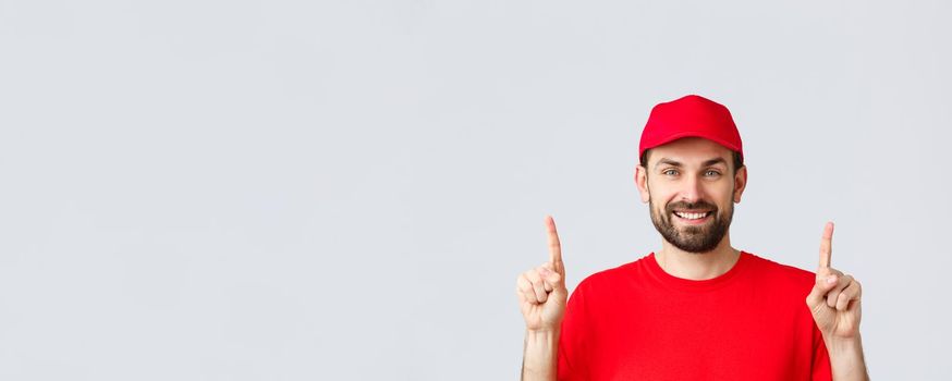 Online shopping, delivery during quarantine and takeaway concept. Cheerful bearded smiling courier in red uniform cap and t-shirt, invite take look at promo, pointing fingers up, grey background.