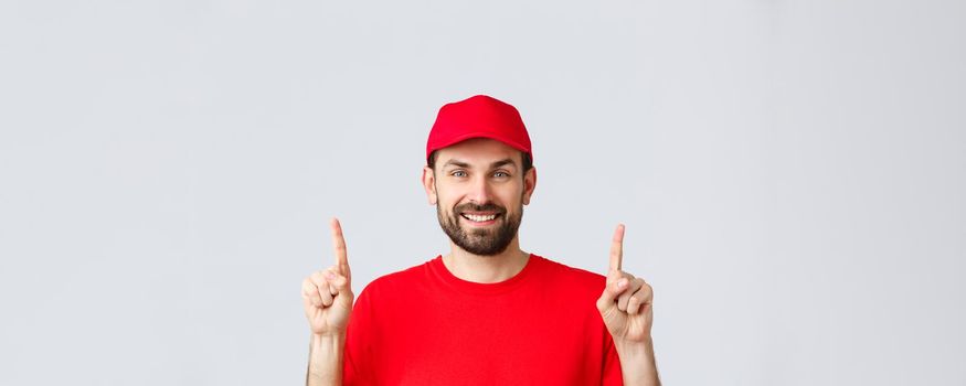Online shopping, delivery during quarantine and takeaway concept. Cheerful bearded smiling courier in red uniform cap and t-shirt, invite take look at promo, pointing fingers up, grey background.